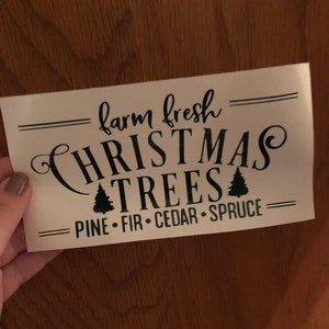 Pu&ograve; includere: Un cartello bianco con testo nero che recita "farm fresh CHRISTMAS TREES PINE - FIR - CEDAR - SPRUCE". Il cartello ha due piccoli alberi di Natale neri.