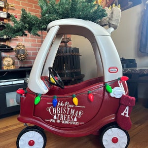 Pu&ograve; includere: Un'auto giocattolo rossa con un tetto bianco e un albero di Natale in cima. L'auto ha un cartello sul lato che dice "Lake Miner Christmas Trees - Pine - Fir - Cedar - Spruce". Ci sono luci colorate infilate intorno all'auto e una calza rossa con la lettera "A" sopra.