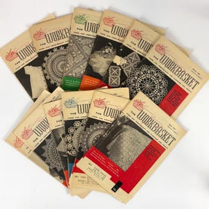 May include: A collection of vintage "Workbasket" magazines, featuring various covers with illustrations of needlework and home crafts. The magazines have a cream-colored background with black and red text and illustrations. The magazines are from 1957.