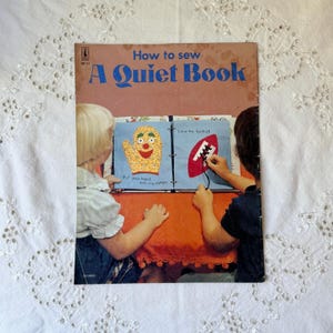 May include: A How to sew A Quiet Book instructional manual with a tan cover. The cover features a photograph of two children interacting with a fabric book showing a yellow mitten graphic and a red football graphic with black lacing.