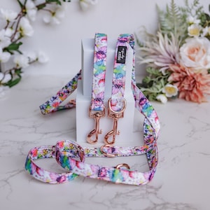 May include: Two floral dog leads with rose gold hardware. The leads are white with a pink, purple and green floral pattern. The leads are on a white surface with a bouquet of flowers in the background.