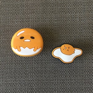 May include: Two yellow and white cartoon egg-shaped charms. One charm is a simple egg with a face, the other is a cartoon character with a face and arms.