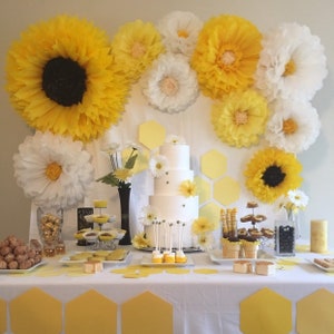 May include: A decorated table with a three-tiered white cake, yellow and white paper flower decorations, and various desserts. The theme is a bee-themed party, with honeycomb decorations and yellow accents.