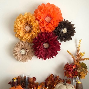 May include: A collection of six tissue paper flowers in shades of orange, yellow, brown, and red. The flowers are arranged on a white wall with a garland of fall leaves and a woven pumpkin in the foreground.