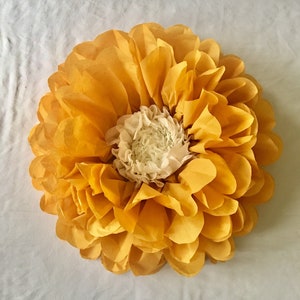 May include: A large, handmade paper flower with layers of yellow petals surrounding a white center. The flower is round and appears to be made of tissue paper. The petals are a bright, sunny yellow, and the center is a soft white.