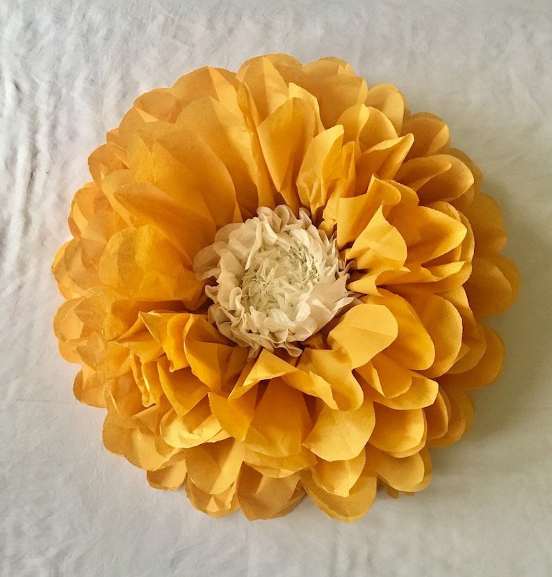 May include: A large, handmade paper flower with layers of yellow petals surrounding a white center. The flower is round and appears to be made of tissue paper. The petals are a bright, sunny yellow, and the center is a soft white.