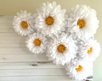 May include: A cluster of white tissue paper flowers with yellow centers, arranged on a white wood background. The flowers are large and fluffy, creating a whimsical and festive atmosphere.