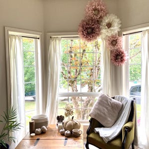May include: A cosy living room with a large window, a wooden coffee table, and a green armchair with a white throw blanket. The window is decorated with large paper flowers in shades of pink and white.