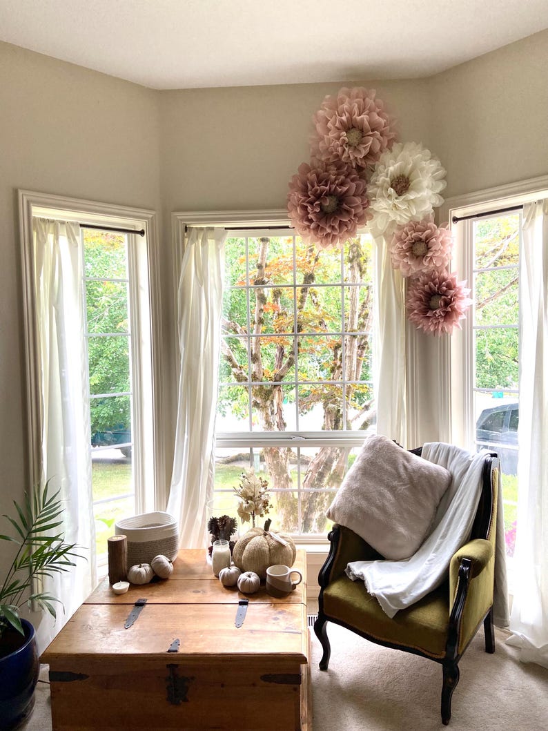 May include: A cosy living room with a large window, a wooden coffee table, and a green armchair with a white throw blanket. The window is decorated with large paper flowers in shades of pink and white.
