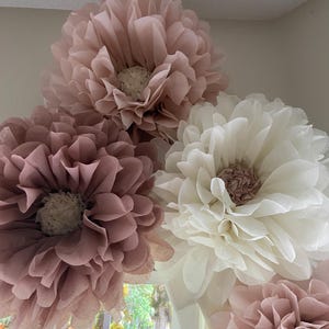 May include: Four large paper flowers in shades of pink and white. The flowers are made of tissue paper and have a ruffled texture. They are arranged in a cluster, with the largest flower at the top.