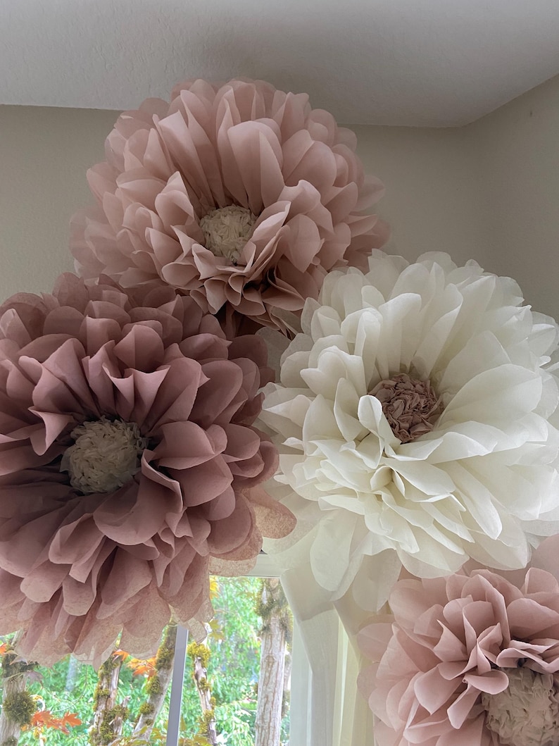 May include: Four large paper flowers in shades of pink and white. The flowers are made of tissue paper and have a ruffled texture. They are arranged in a cluster, with the largest flower at the top.