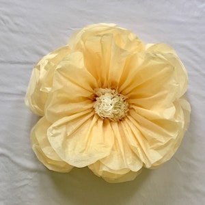 May include: A large, handmade paper flower in shades of pale yellow and cream. The flower has multiple layers of petals, with a textured center. The petals are ruffled, creating a soft, delicate appearance. The flower is likely used for decoration.