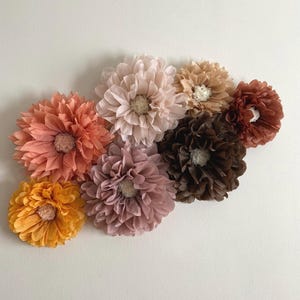 May include: An assortment of handmade paper flowers in various colors, including peach, pink, brown, and yellow. The flowers are arranged in a cluster, with detailed petal textures and a central, textured core. The flowers are likely for home decor.