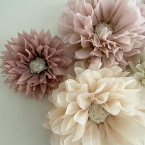 May include: Three large paper flowers in shades of pink and cream. The flowers are made of tissue paper and have a layered, ruffled appearance.