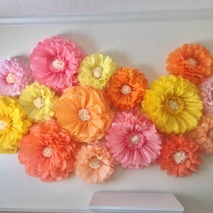 Puede incluir: Un conjunto de grandes flores de papel de colores en tonos rosa, amarillo, naranja y melocot&oacute;n, montadas en una pared blanca. Las flores tienen un dise&ntilde;o de p&eacute;talos en capas y un centro texturizado, creando una exhibici&oacute;n decorativa.
