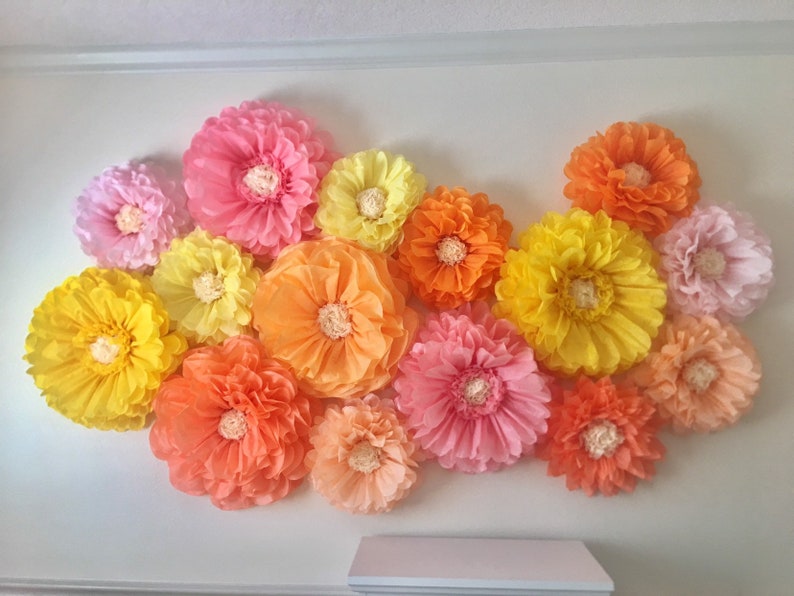 Puede incluir: Un conjunto de grandes flores de papel de colores en tonos rosa, amarillo, naranja y melocot&oacute;n, montadas en una pared blanca. Las flores tienen un dise&ntilde;o de p&eacute;talos en capas y un centro texturizado, creando una exhibici&oacute;n decorativa.