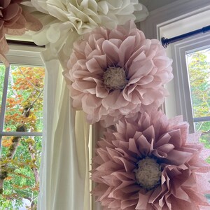 May include: Four large tissue paper flowers in shades of pink and white are hanging from a white ceiling. The flowers are arranged in a corner of a room, with the largest flower at the top and the smallest flower at the bottom.