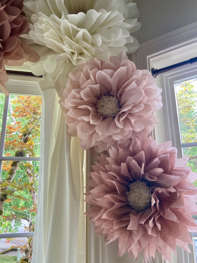 May include: Four large tissue paper flowers in shades of pink and white are hanging from a white ceiling. The flowers are arranged in a corner of a room, with the largest flower at the top and the smallest flower at the bottom.