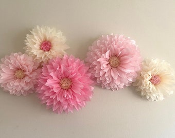 Könnte beinhalten: Fünf große Papierblumen in Rosa- und Cremetönen. Die Blumen sind in einer Reihe an einer weißen Wand angeordnet.