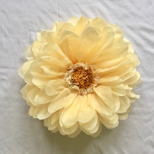 May include: A large, handmade paper flower in shades of pale yellow and cream. The flower has multiple layers of petals, creating a full, rounded shape. The center of the flower is a darker shade of yellow and cream.