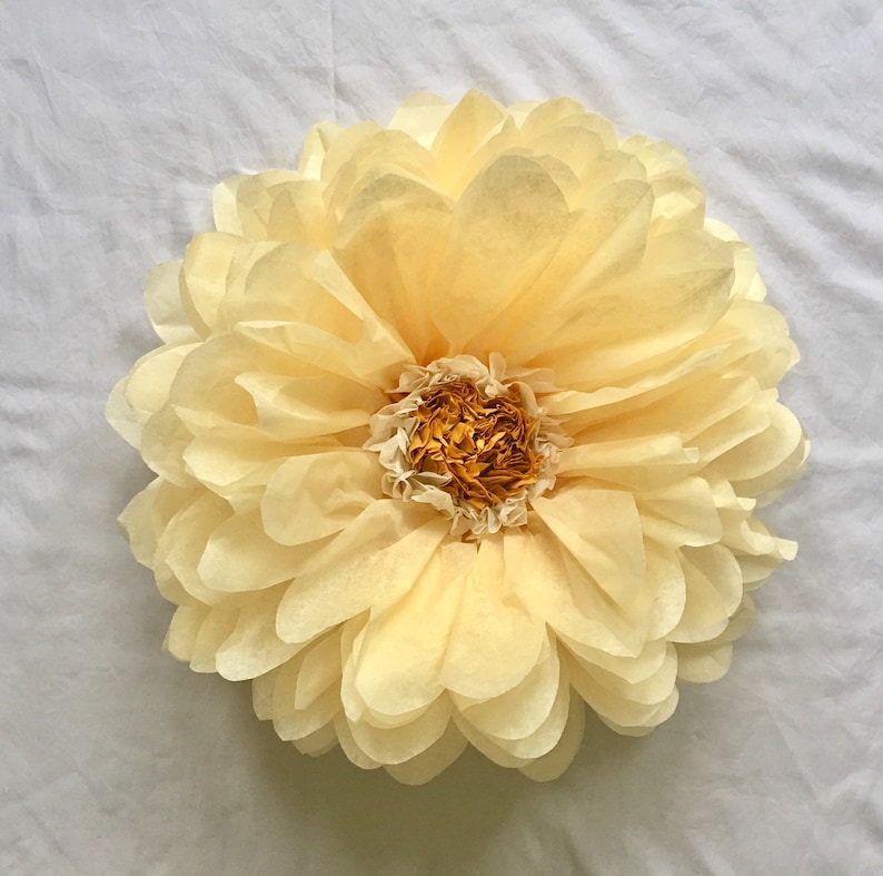 May include: A large, handmade paper flower in shades of pale yellow and cream. The flower has multiple layers of petals, creating a full, rounded shape. The center of the flower is a darker shade of yellow and cream.