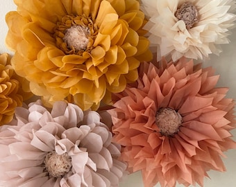 May include: Five tissue paper flowers in various shades of pink, peach, and yellow. The flowers are arranged on a white wall.