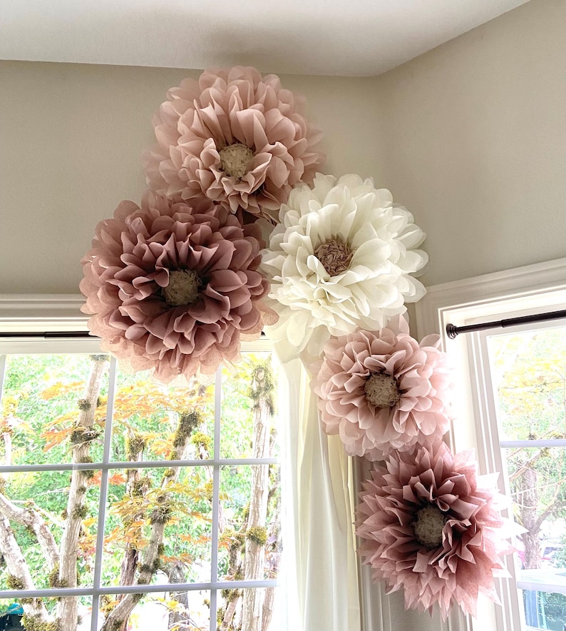 May include: Five large paper flowers in shades of pink and white are arranged in a corner of a room. The flowers are made of tissue paper and have a delicate, ruffled appearance.