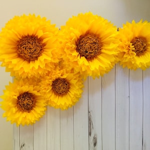 May include: A collection of five large yellow paper sunflowers arranged in a row on a white wood background.