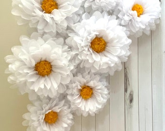 May include: White paper flowers with yellow centers are arranged on a white wooden background. The flowers are large and fluffy, resembling daisies. The arrangement is a decorative accent.
