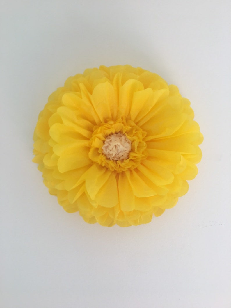 Puede incluir: Una flor de papel amarilla, grande y redonda, con m&uacute;ltiples capas de p&eacute;talos. El centro de la flor es de un color beige m&aacute;s claro. La flor est&aacute; sobre un fondo blanco liso.