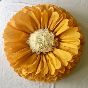 May include: A large, round decorative paper flower with layers of yellow petals surrounding a cream-colored center. The petals are arranged in a circular pattern, resembling a giant daisy or sunflower. The flower is made of delicate paper.