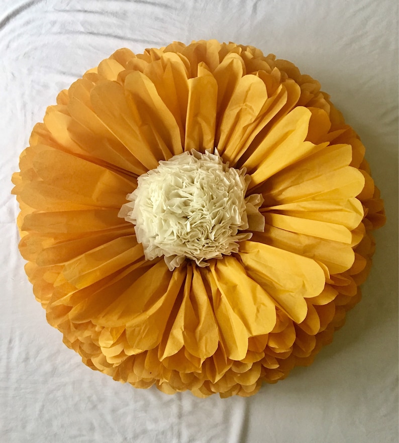 May include: A large, round decorative paper flower with layers of yellow petals surrounding a cream-colored center. The petals are arranged in a circular pattern, resembling a giant daisy or sunflower. The flower is made of delicate paper.