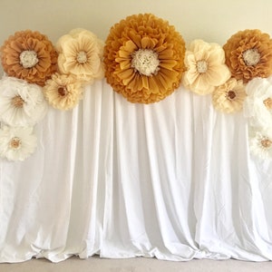 May include: A backdrop featuring large paper flowers in shades of gold, cream, and white. The flowers are arranged above a white fabric curtain, creating a decorative display. The backdrop is suitable for events and photo shoots.