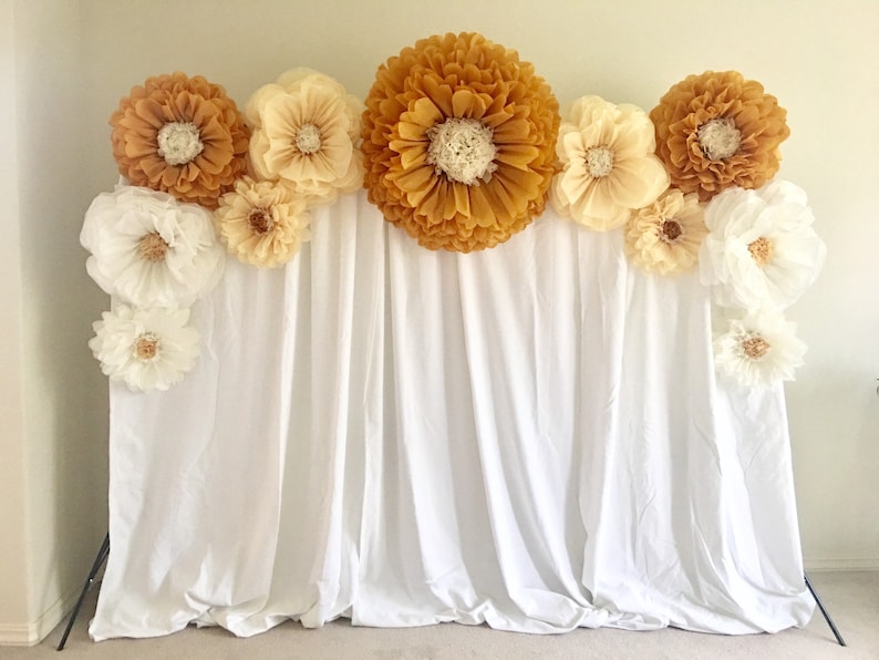 May include: A backdrop featuring large paper flowers in shades of gold, cream, and white. The flowers are arranged above a white fabric curtain, creating a decorative display. The backdrop is suitable for events and photo shoots.