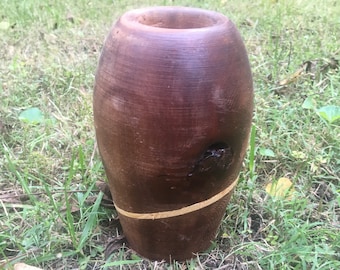 May include: A brown wooden vase with a light brown band around the middle. The vase is sitting on a bed of green grass.