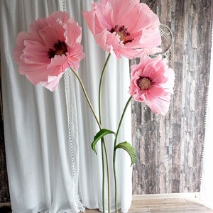 以下が含まれることがあります： 緑色の茎と緑色の金属製の土台に、大きなピンク色の紙の花が3つあります。花は花瓶のような形に配置されています。