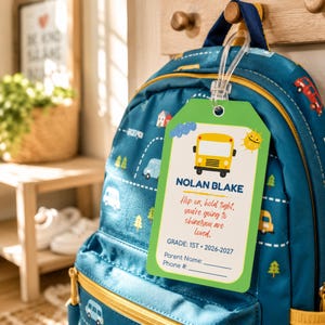 May include: A rectangular green luggage tag with a yellow school bus graphic, the name NOLAN BLAKE, and the text: Hop on, hold tight, you're going to shine! you are loved. GRADE: 1ST • 2026-2027, Parent Name: ____, Phone #: ____, attached to a blue backpack.