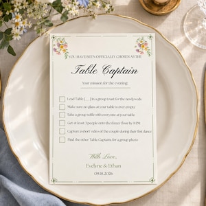 Könnte beinhalten: Eine rechteckige Hochzeitstischkarte mit cremefarbenem Hintergrund, grünem Rand und Blumenmotiven. Sie enthält den Text: YOU HAVE BEEN OFFICIALLY CHOSEN AS THE Table Captain. Ihre Mission für den Abend umfasst sechs Aufgaben, signiert mit With Love, Evelyne & Ethan 18.09.2026.