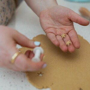 May include: Close-up of hands holding star-shaped cookie cutouts and a white cookie cutter. A sheet of brown dough has star cutouts. The scene suggests baking or crafting, with a focus on the star shapes.