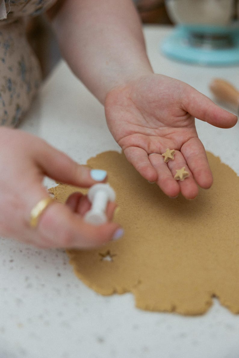 May include: Close-up of hands holding star-shaped cookie cutouts and a white cookie cutter. A sheet of brown dough has star cutouts. The scene suggests baking or crafting, with a focus on the star shapes.