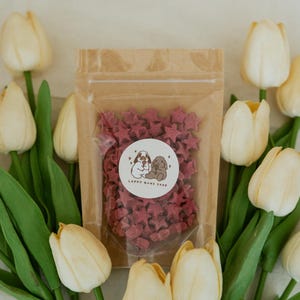 May include: A clear bag filled with star-shaped treats, with a sticker that reads "LOPPY BUNE SHOP". The bag is surrounded by white and yellow tulips with green stems and leaves. The treats are a deep red color.