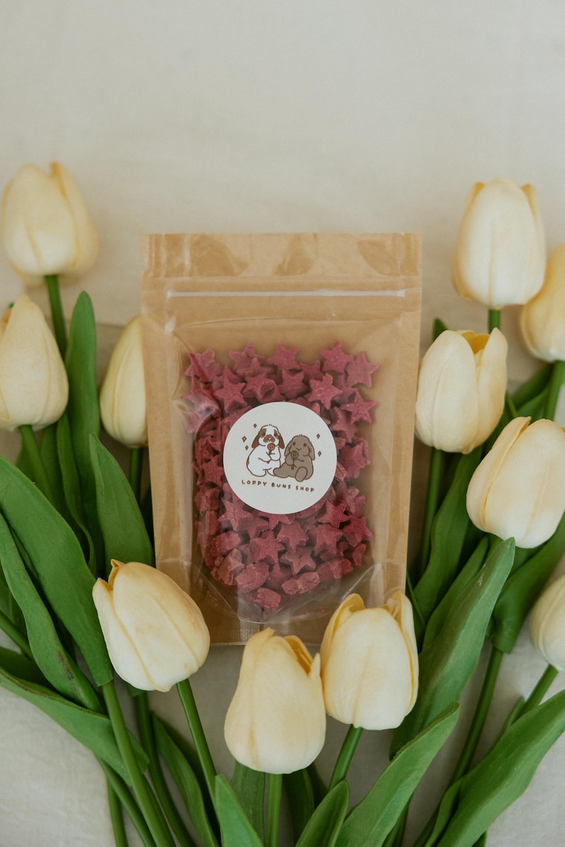 May include: A clear bag filled with star-shaped treats, with a sticker that reads "LOPPY BUNE SHOP". The bag is surrounded by white and yellow tulips with green stems and leaves. The treats are a deep red color.