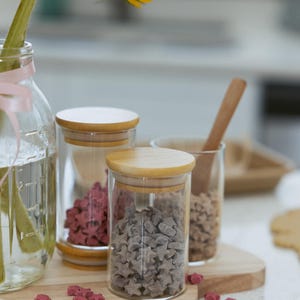 May include: A clear glass jar with a wooden lid contains gray star-shaped candies. Another jar holds pink star-shaped candies. A sunflower in a glass jar with a pink ribbon is in the background. The jars sit on a wooden cutting board.