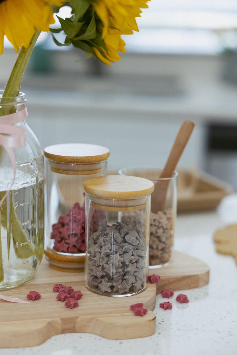 May include: A clear glass jar with a wooden lid contains gray star-shaped candies. Another jar holds pink star-shaped candies. A sunflower in a glass jar with a pink ribbon is in the background. The jars sit on a wooden cutting board.