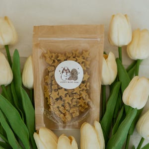 May include: A clear plastic bag filled with star-shaped dog treats, with a circular sticker that reads "Lappy Buns Shop". The bag is surrounded by white tulips with green stems and leaves. The treats are light brown.