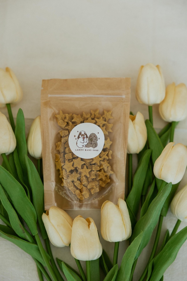 May include: A clear plastic bag filled with star-shaped dog treats, with a circular sticker that reads "Lappy Buns Shop". The bag is surrounded by white tulips with green stems and leaves. The treats are light brown.