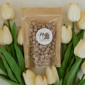 May include: A clear plastic bag filled with star-shaped treats, with a sticker that says "Loppy Buns Shop". The bag is surrounded by cream-colored tulips with green stems and leaves.