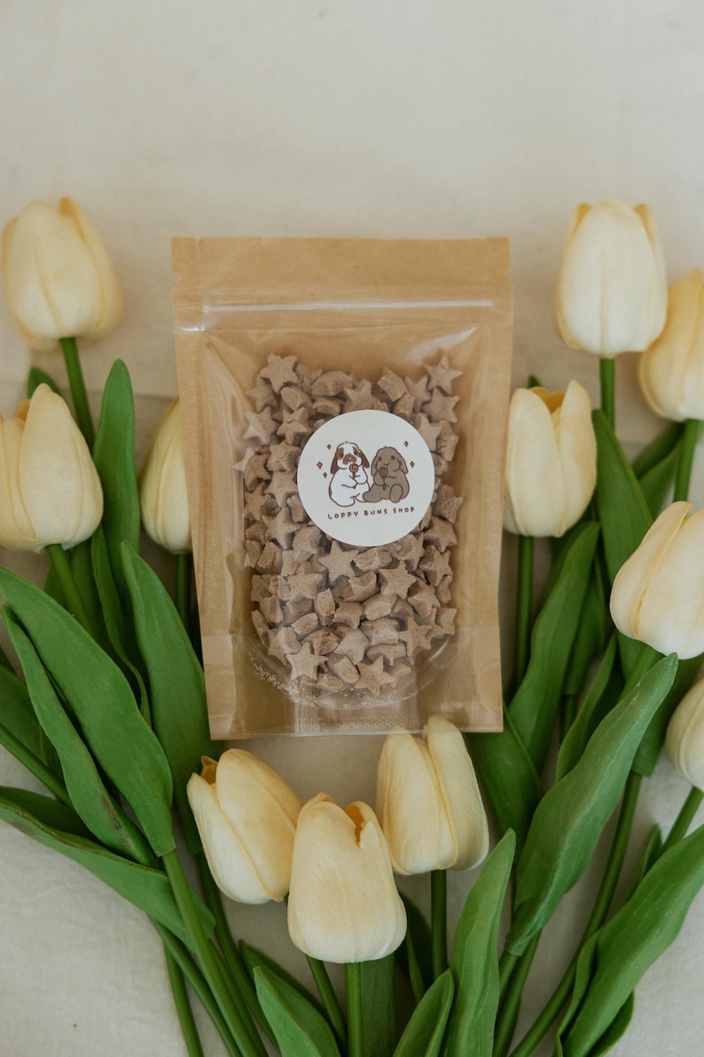 May include: A clear plastic bag filled with star-shaped treats, with a sticker that says "Loppy Buns Shop". The bag is surrounded by cream-colored tulips with green stems and leaves.