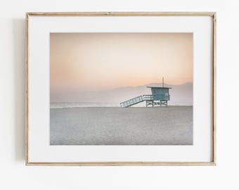 May include: A framed wall art print featuring a light blue lifeguard tower standing on a sandy beach at sunset. The scene includes a calm ocean horizon under a soft, hazy peach and cream sky, all enclosed within a simple wooden frame.