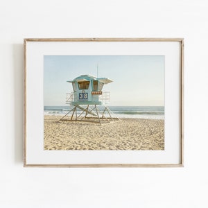 May include: A framed wall art print featuring a light blue lifeguard tower on a sandy beach. The tower is elevated on a metal frame and displays the number 38 on its front panel. The ocean and horizon are visible in the background under a clear sky.